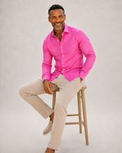 Load image into Gallery viewer, Man wearing a bright pink shirt and beige pants sitting on a wooden stool against a plain background