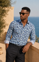 Load image into Gallery viewer, Man wearing a blue floral shirt and sunglasses, standing by a scenic coastal area.