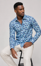 Load image into Gallery viewer, Man wearing a blue floral shirt sitting on a stool against a gray background