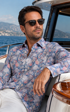 Load image into Gallery viewer, A man wearing a blue and pink floral long-sleeve button-up shirt and white trousers, sitting on a yacht with the sea and mountains in the background.