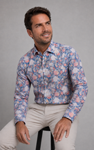 Load image into Gallery viewer, A man wearing a long-sleeve button-up shirt with a blue, pink, and white floral pattern, paired with beige trousers, sitting on a stool against a gray studio background.