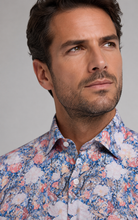 Load image into Gallery viewer, A close-up of a man wearing a button-down shirt with a dense floral pattern in blue, pink, and white, set against a plain gray studio background.