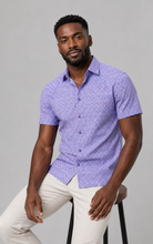 Load image into Gallery viewer, A man wearing a short-sleeve button-up shirt with a purple and white geometric pattern, paired with off-white trousers, sitting on a stool against a solid gray background.